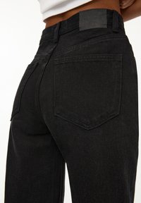 Gros plan sur une personne portant un jean en denim noir taille haute avec des poches arrière et un patch en cuir de marque à la ceinture.