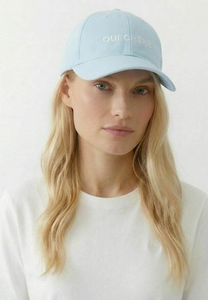 Blonde Frau mit offenem Haar trägt eine hellblaue Kappe mit der Aufschrift "OUI CHÉRIE" und ein weißes Hemd mit kleinem Herzdetail auf der Brust.
