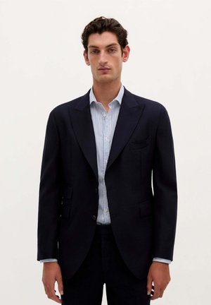 Blazer marino con solapas de muesca, dos bolsillos frontales y un ajuste entallado, combinado con una camisa de rayas azul claro y pantalones oscuros.