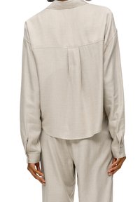 Light beige long-sleeve blouse with a collar, featuring a back pleat and buttoned cuffs, made from a soft, textured fabric.