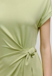 Hellgrünes Oberteil aus weichem Stoff, mit einem verdrehten Vorderdesign und einem geknoteten Detail an der Taille sowie umgeschlagenen kurzen Ärmeln.