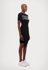Čierne krátke šaty s potlačou "VERSACE JEAN COUTURE". Modelka ich dopĺňa bielymi teniskami, stojí na neutrálnom pozadí.