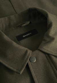 Olīvu zaļa jaka ar mīkstu audumu pie apkakles un matētu melnu pogu. Satur birku ar zīmola nosaukumu "GABBA" identifikācijai.