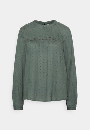 Blouse verte semi-transparente à manches longues avec texture à pois, bordure en dentelle sur la poitrine, col rond et fermeture à boutons dans le dos.