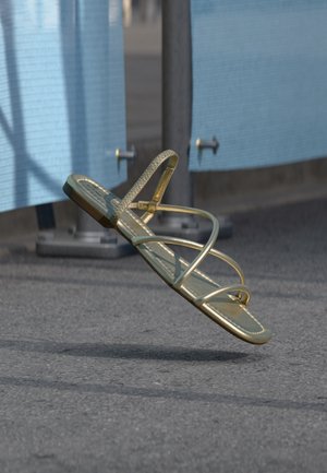 Złoty, pojedynczy sandał na cienkich paskach zawieszony w powietrzu nad asfaltem, w tle niebieskie tkaniny i metalowe słupy.
