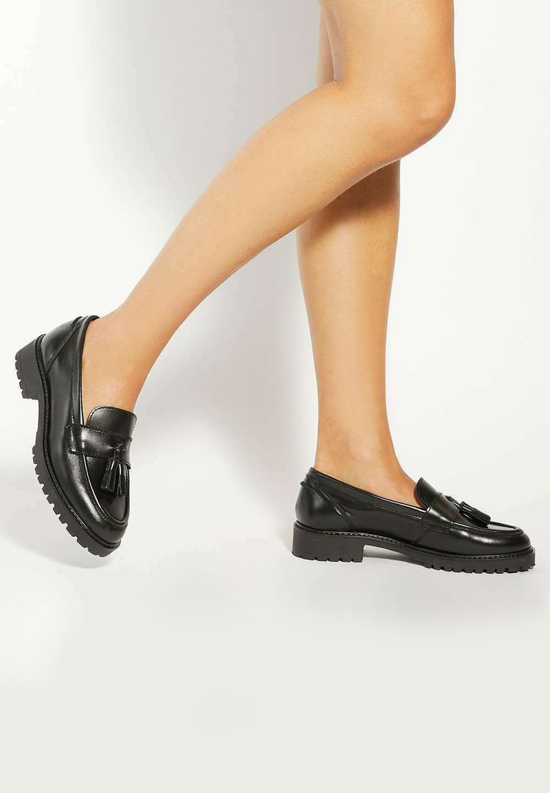Black leather loafers with a tassel detail, low chunky sole, and smooth finish. Side view showcases a rounded toe and sleek design.
