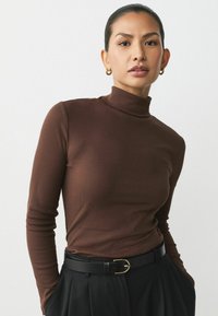 Brown ribbed turtleneck top with long sleeves. Paired with black high-waisted trousers and a gold-buckled black belt.