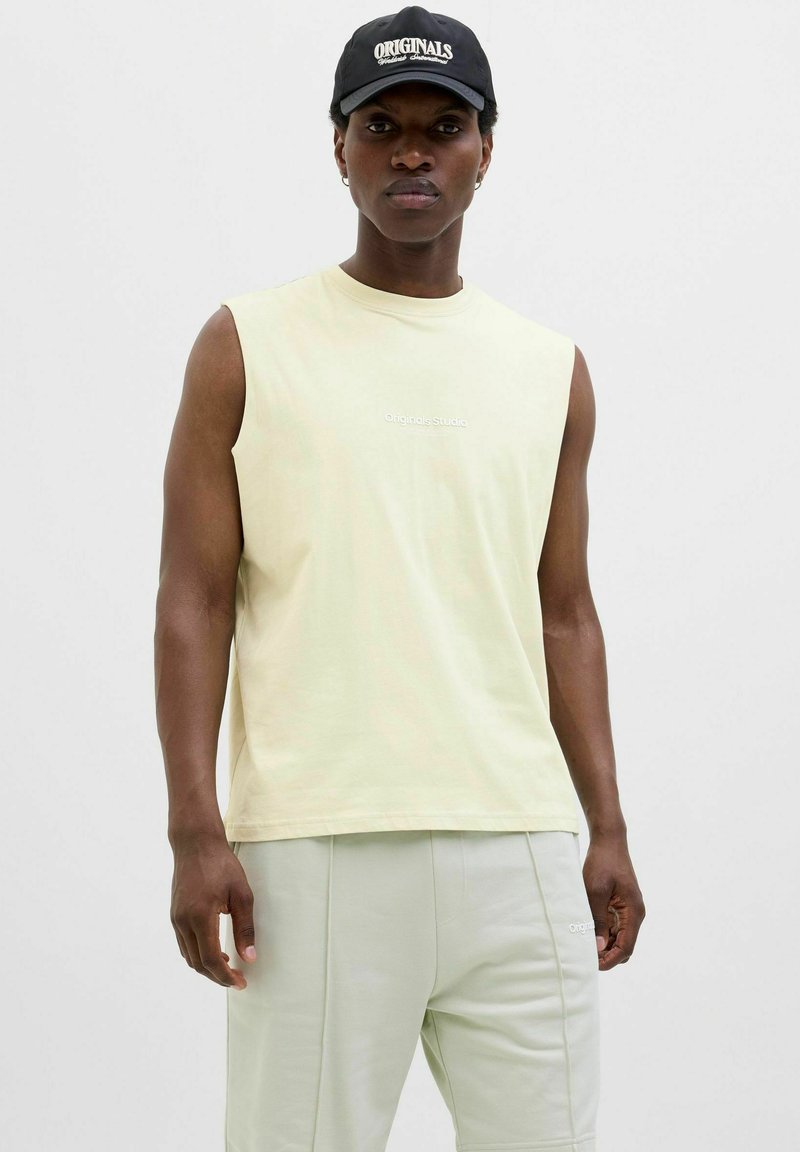 T-shirt sans manches jaune clair avec le texte "Originals Studio". Matière en coton lisse. Associé à un pantalon vert clair, le tout dans une coupe décontractée et confortable.