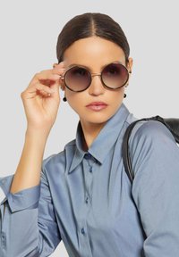 Runde Sonnenbrillen mit dunklen Gläsern und einem dünnen goldenen Rahmen. Das Modell trägt ein hellblaues Hemd mit Knopfleiste und hält die Sonnenbrille mit einer Hand.