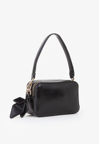 Borsa a spalla rettangolare nera con zip dorata, manico corto e etichette decorative in pelle attaccate su sfondo bianco.