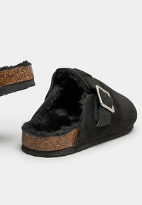 Zapatillas de ante negro sin cordones con forro de pelo, plantilla de corcho y un acento de hebilla plateada. Suela plana de goma con fondo texturizado.