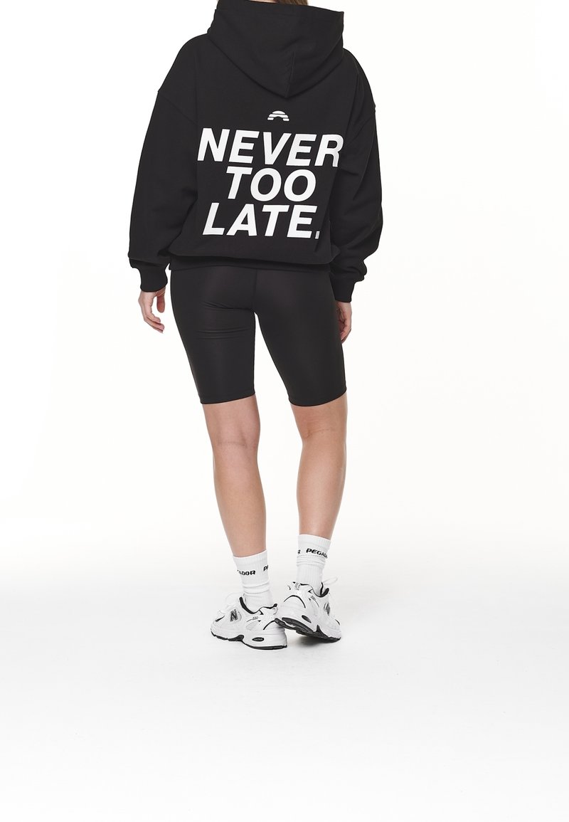Personne portant un sweat à capuche noir avec le texte "NEVER TOO LATE.", un short de cyclisme noir, des chaussettes blanches et des baskets blanches, debout de dos.