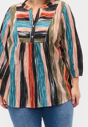 Personne grande taille portant un chemisier à manches longues rayé multicolore avec des boutons et un jean bleu, debout devant un fond uni.