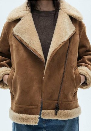 Braune Wildleder-Shearlingjacke mit cremefarbenem Fleece-Futter, getragen über einem dunkelbraunen Pullover und blauen Jeans, Hände in den Jackentaschen.