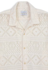 Camicia con bottoni in pizzo di color crema chiaro, con intricati motivi a diamante e floreali, dal taglio rilassato e dalla leggera consistenza. Dettagli dei bottoni visibili.