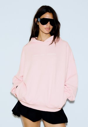 Femme portant un sweat à capuche rose pâle oversize et de grandes lunettes de soleil noires, les mains dans les poches et une jupe noire sur un fond blanc uni.