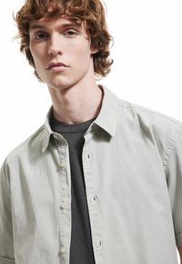 Camisa de botón gris claro con cuello, usada sobre un suéter gris oscuro. Textura suave, detalles mínimos y botones visibles en la parte frontal.