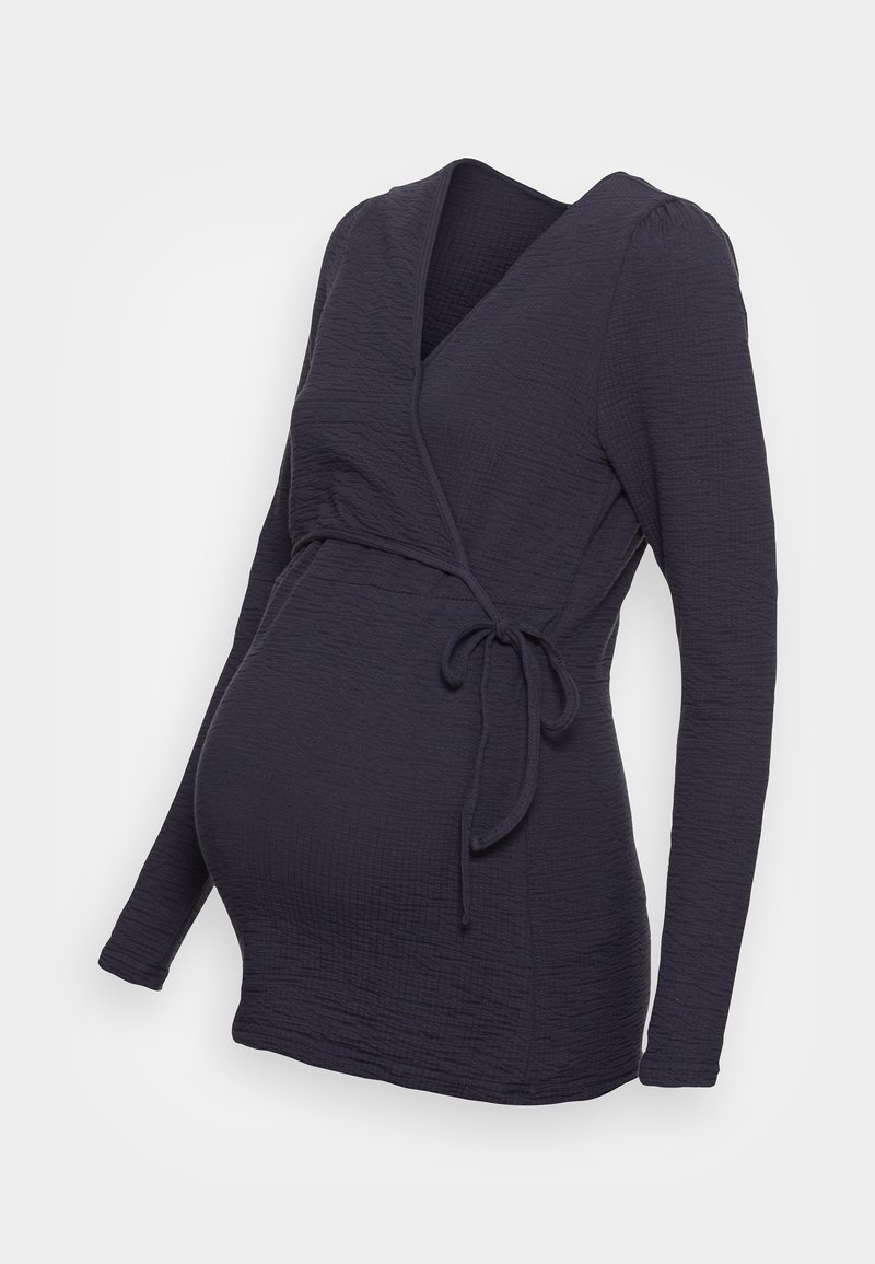 Haut de maternité à manches longues en voile bleu marine avec un tissu texturé et un lien sur le côté à la taille, conçu pour un ajustement confortable sur un ventre de grossesse.