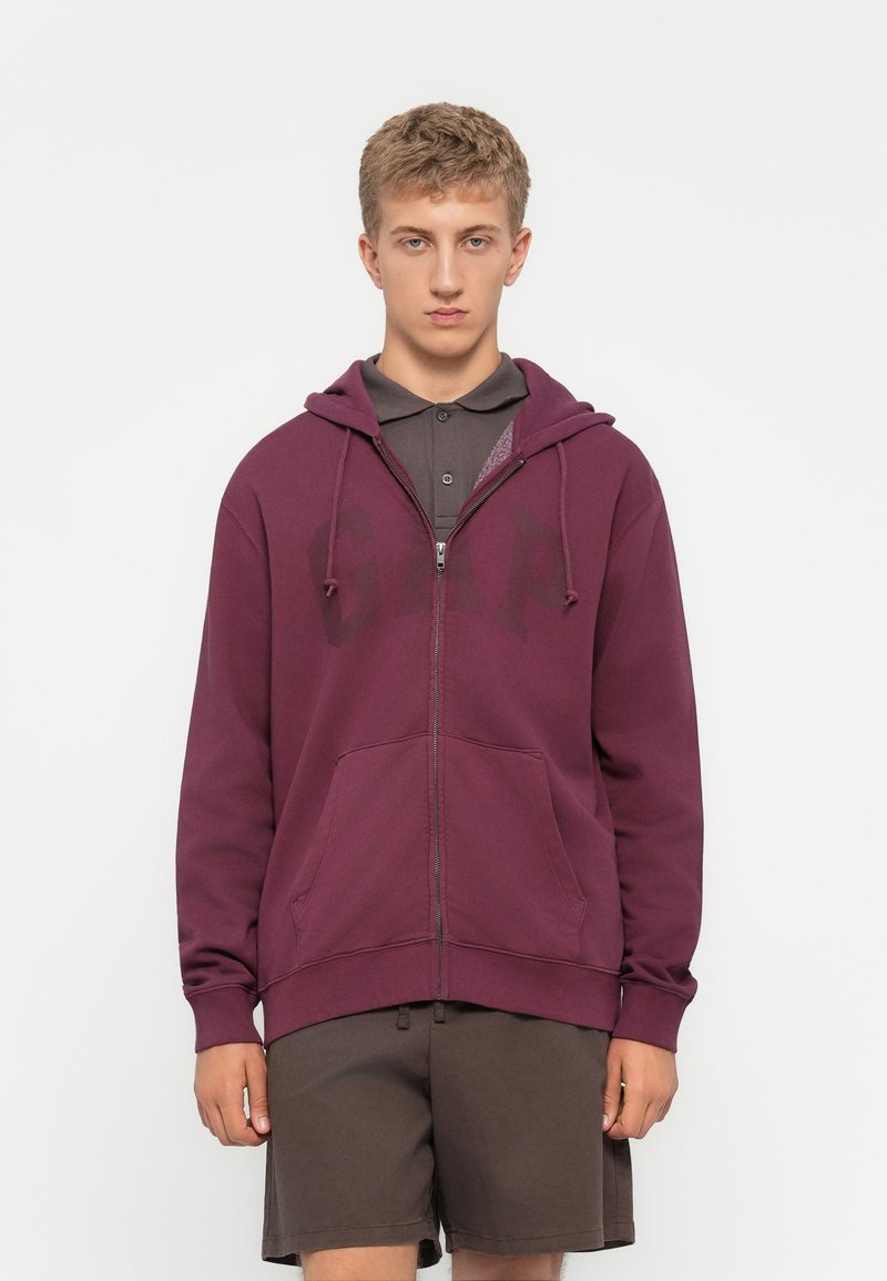 Ung mand iført en maroon zip-up hættejakke med "GAP" tekst og brune shorts, stående foran en helt hvid baggrund.