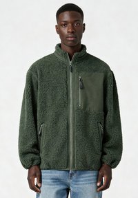 Veste en polaire verte avec une texture moelleuse, fermeture éclair à l'avant et poches latérales. Comprend une poche poitrine en tissu uni et des poignets côtelés.