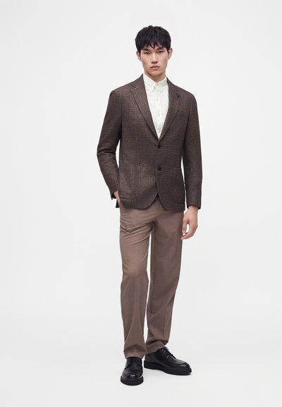 Homme portant un blazer marron texturé, une chemise blanche boutonnée, un pantalon taupe et des chaussures habillées noires, debout devant un fond blanc.