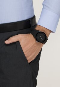 Black rubber watch with a round face and minimal markings, worn on a wrist, alongside a blue long-sleeve shirt and dark trousers.