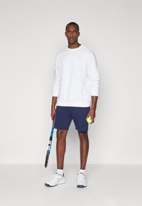 Sweatshirt blanc pour homme, shorts navy, baskets blanches. Tenant une raquette de tennis et une balle verte. Design simple, en mélange de coton, coupe athlétique.