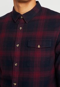 Primo piano di una persona che indossa una camicia a quadretti bordeaux scuro e blu navy con taschino sul petto abbottonato.