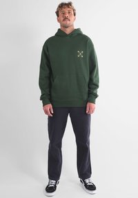 Sweat à capuche vert en polaire avec une poche frontale, présentant un petit motif graphique sur le côté gauche de la poitrine et des poignets côtelés.