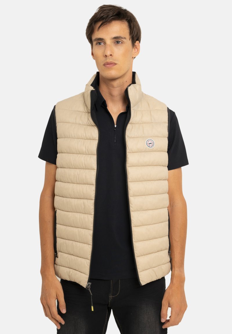 Veste matelassée beige avec coutures horizontales, col haut et fermeture zippée. Comprend un patch logo et est portée sur une chemise noire.