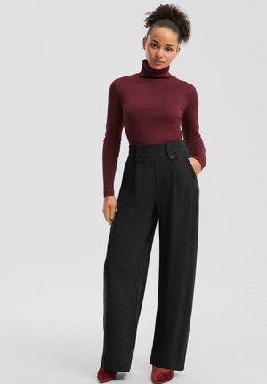 Pullover mit Stehkragen in Bordeaux, taillierte Passform. Schwarze, hochgeschnittene Hosen mit weitem Bein, Doppelknebel-Details und Seitentaschen. Rote Schuhe.