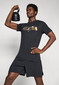 Sort Nike T-shirt med "JUST DO IT." i levende farver, parret med sorte shorts. Personen løfter en sort kettlebell. Enkel baggrund.