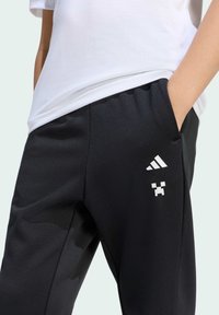Černé joggingové kalhoty vyrobené z hladké tkaniny. Mají bílé logo Adidas a pixelový design na noze, s bočními kapsami a elastickým pasem.