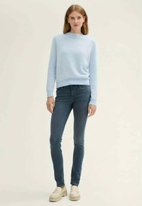 Sieviete, ģērbusies gaiši zilā svītrainā džemperī, tumšās šaurajās džinsi un krēmkrāsu platformas mokasīnos, stāv uz vienkārša bēša fona.