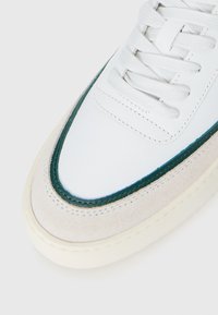 Baskets blanches avec une bordure verte, un détail en daim beige, des lacets blancs et une semelle écrue, présentées en gros plan sur la zone des orteils.