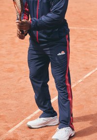 Marina atletiska byxor med röda sidsömmar, tillverkade av lätt material. Bärs med vita sneakers på en lergolvtennisbana.