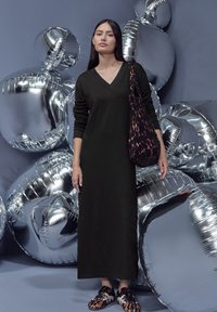 Schwarzes langärmeliges Kleid mit V-Ausschnitt und geradem Schnitt. Kombiniert mit einer Schultertasche im Leopardenmuster und gemusterten Ballerinas, vor einem Hintergrund aus metallischen Luftballons.