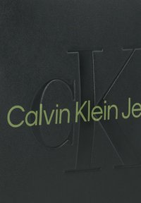 Schwarzes strukturiertes Material mit geprägtem "CK"-Logo und "Calvin Klein Jeans" in fettem grünen Schriftzug. Sauberes, modernes Design.