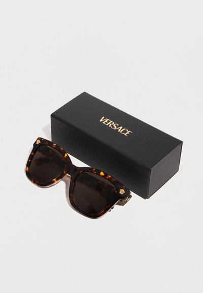 Lunettes de soleil carrées écaille de tortue avec des verres foncés à côté d'une boîte noire ornée du logo doré "Versace" sur une surface blanche.