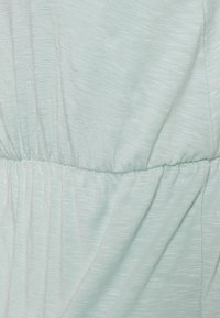 Tecido verde claro com uma superfície suave e texturada. Inclui uma seção franzida ao longo da cintura, criando um design fluido.