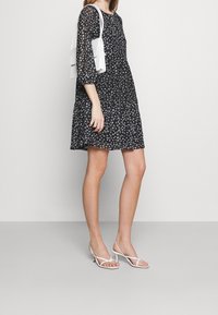 Kvinna iförd en svart blommig klänning, vita strap-on kitten heels och bär en vit axelremsväska mot en enkel bakgrund.