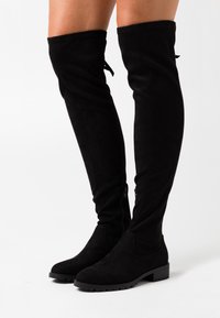Bottes noires en daim montant au-dessus du genou avec un bout arrondi, un talon bloc et des fermetures éclair latérales. Présentent une texture lisse et un détail de couture minimal.