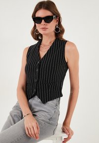 Čierny bezrukávový vest s bielymi prúžkami, jednoradové gombíky a špicatý lem, skombinovaný so šedými jeansami s vysokým pásom a slnečnými okuliarmi.