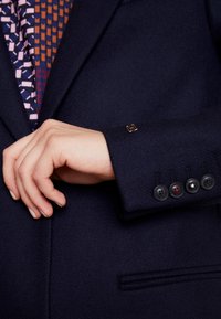 Blazer azul marino oscuro con una tela texturizada, cuatro botones negros con un acento rojo y un detalle de logo dorado en el puño.