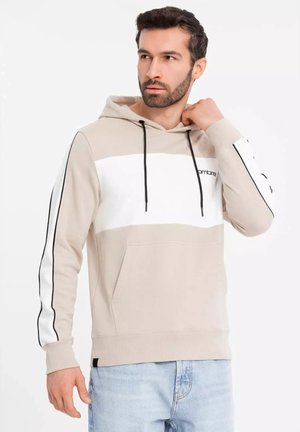 Sudadera beige con una franja horizontal blanca, cordones negros y acentos laterales. Presenta un bolsillo delantero y una textura de tela suave y lisa.