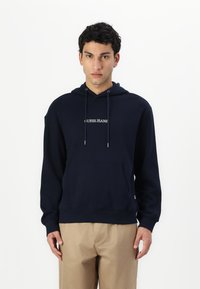 Tengerészeti kapucnis felső húzózsinórral és elülső zsebbel. A mellkason a fehér "GUESS JEANS" logó található. Pamut anyag, laza szabás. Bézs nadrággal kombinálva.