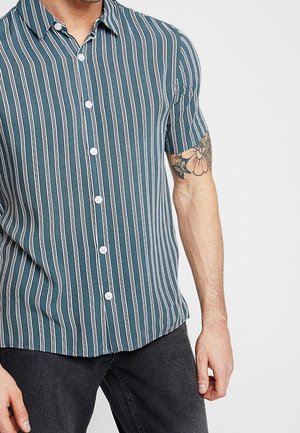 Uomo con camicia a maniche corte color teal con righe verticali bianche, jeans neri e tatuaggio floreale sull'avambraccio sinistro, in piedi davanti a uno sfondo semplice.