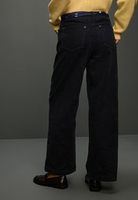 Szerokie spodnie sztruksowe w ciemnym granacie, z teksturowaną, prążkowaną powierzchnią, dwoma tylnymi kieszeniami oraz zapięciem na guzik z wzorzystym paskiem w talii.