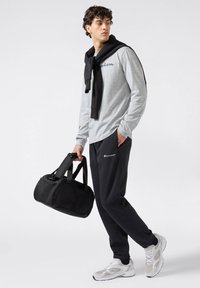 Maglietta a maniche lunghe grigia chiara con logo "Champion", maglione nero a collo giro con le maniche riempite, pantaloni della tuta neri, scarpe sportive grigie e borsa da palestra nera.