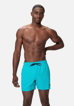 Fit man with short hair wearing bright turquoise swim shorts with black drawstring, standing with one hand behind his back.